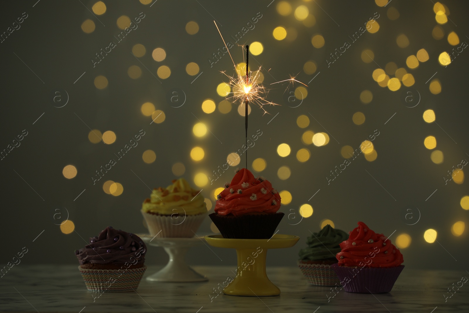 Photo of Different colorful cupcakes and one with sparkler on table against blurred lights