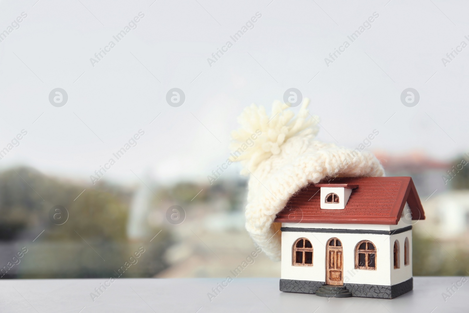 Photo of House model with knitted hat on table against blurred background, space for text. Heating concept