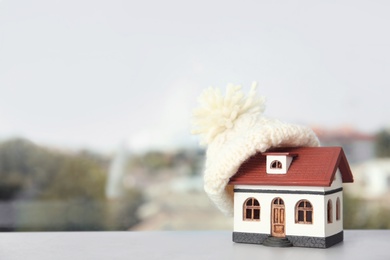 Photo of House model with knitted hat on table against blurred background, space for text. Heating concept