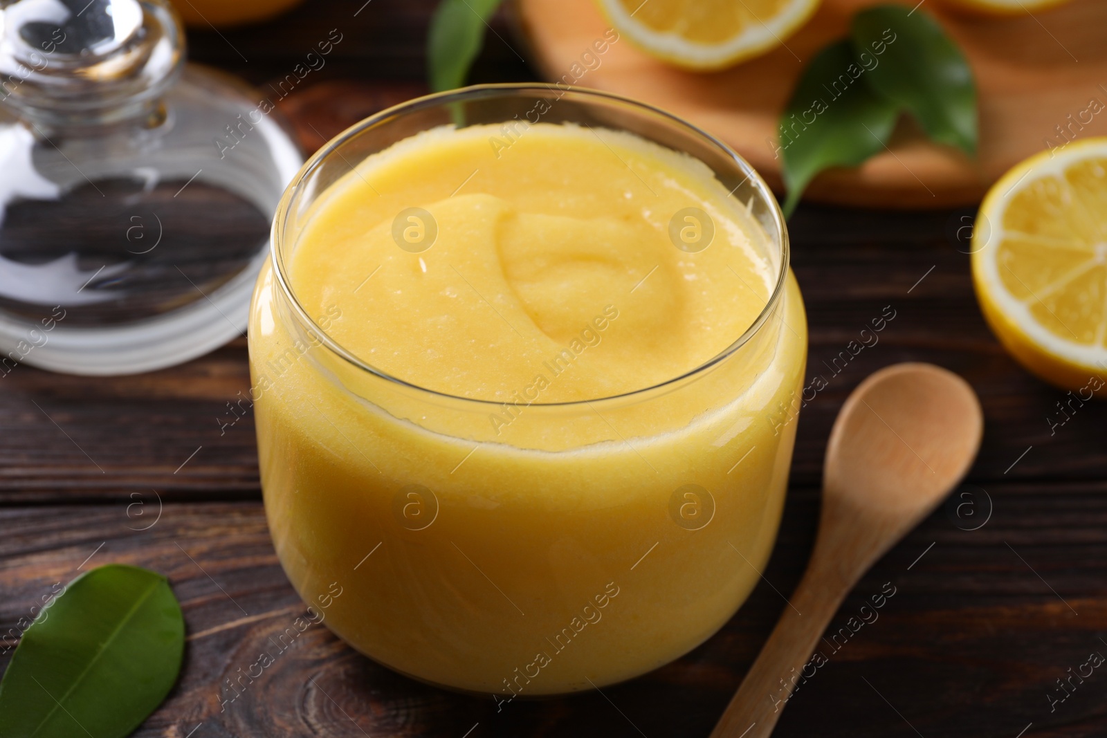 Photo of Delicious lemon curd in glass jar, fresh citrus fruit and spoon on wooden table, closeup