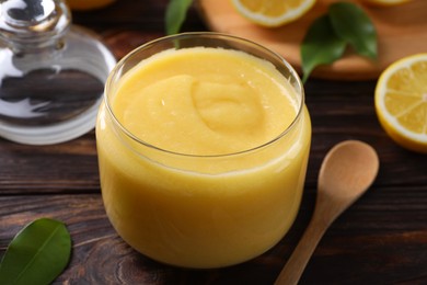 Photo of Delicious lemon curd in glass jar, fresh citrus fruit and spoon on wooden table, closeup