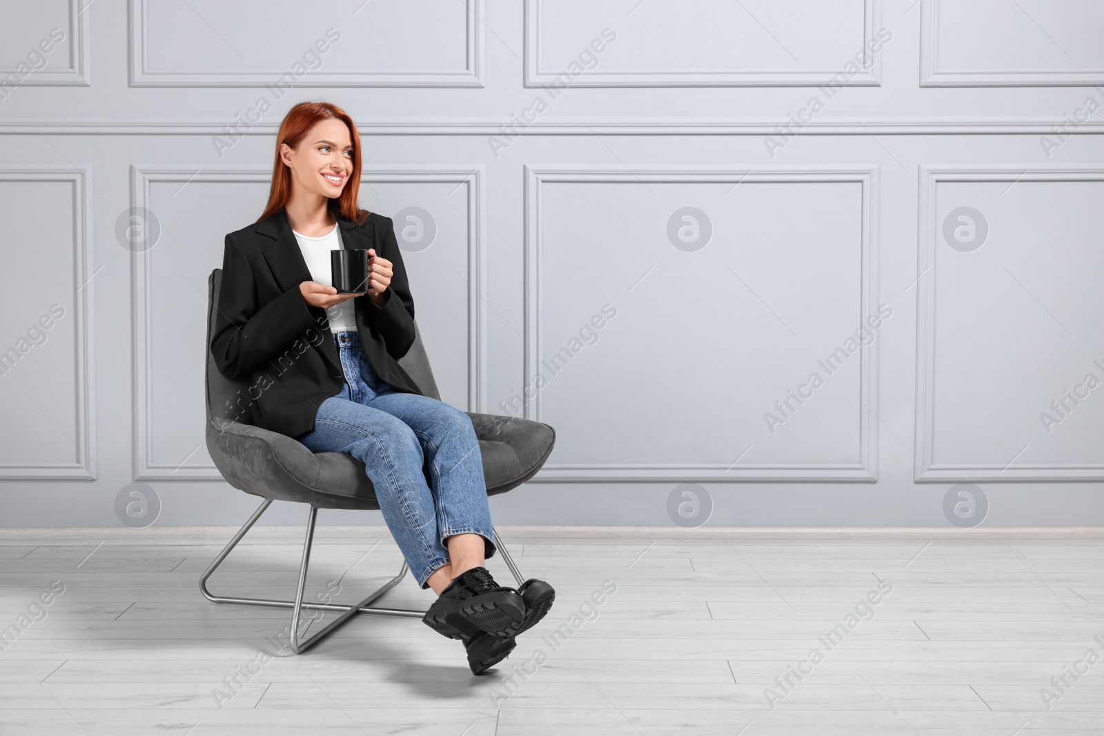 Photo of Happy young woman with cup of drink sitting in armchair near light grey wall indoors. Space for text