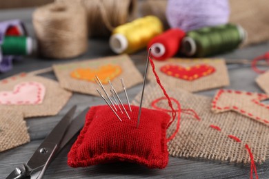 Photo of Pincushion with needles, scissors and pieces of burlap fabric on grey wooden table, closeup