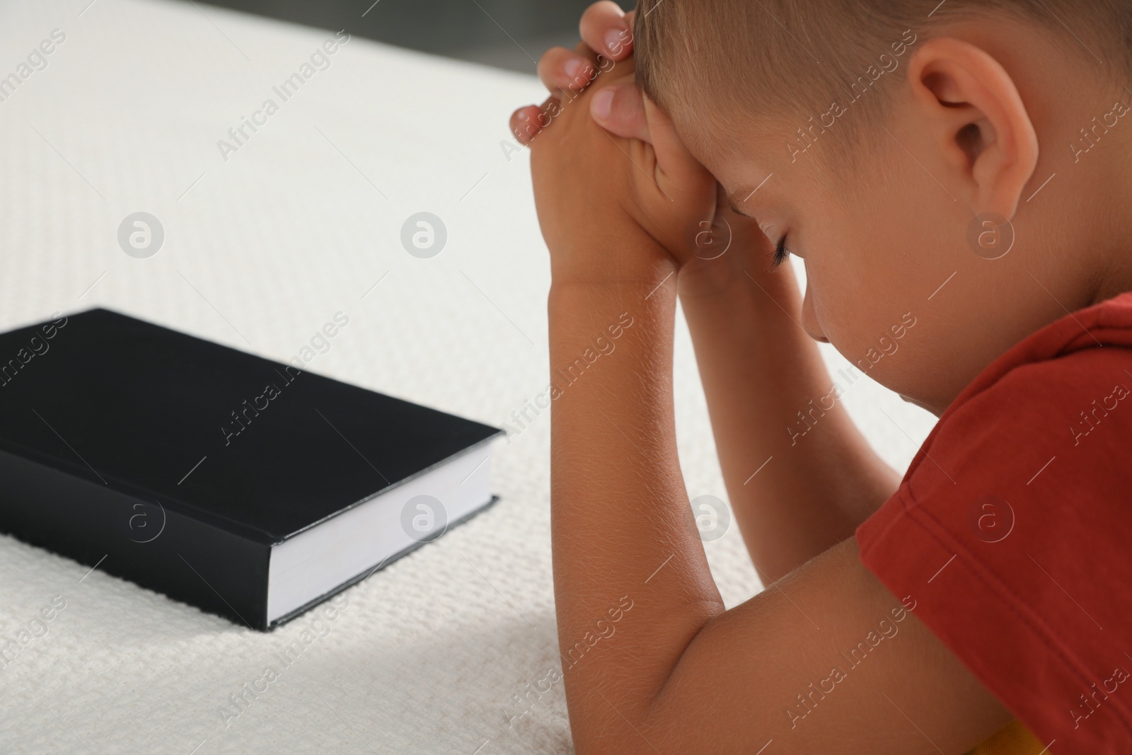 Photo of Cute little boy with hands clasped together saying bedtime prayer over Bible at home, closeup