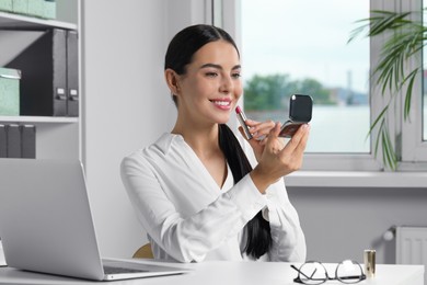 Young woman using cosmetic pocket mirror while applying makeup in office