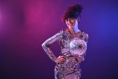 Photo of Beautiful young woman with disco ball posing on color background in neon lights. Space for text