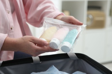 Photo of Woman holding plastic bag of cosmetic travel kit over suitcase indoors, closeup. Bath accessories
