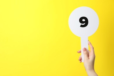 Photo of Woman holding auction paddle with number 9 on yellow background, closeup. Space for text