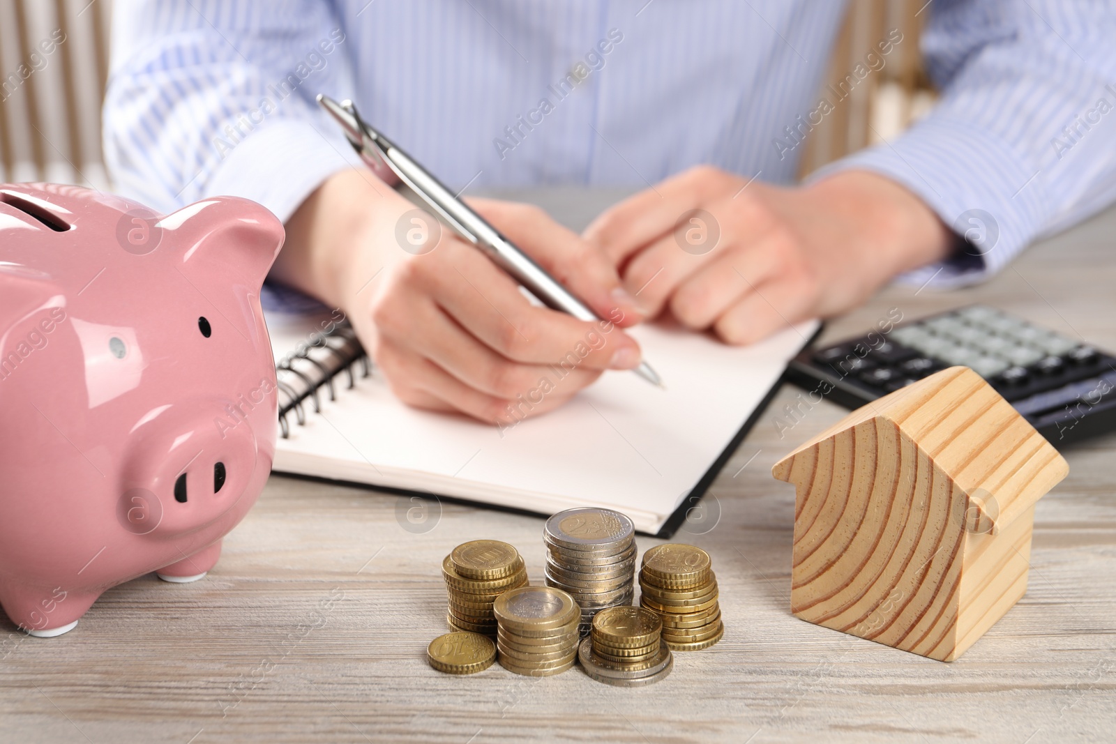 Photo of Woman planning budget at wooden table, focus on house model, coins and piggy bank