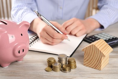 Photo of Woman planning budget at wooden table, focus on house model, coins and piggy bank