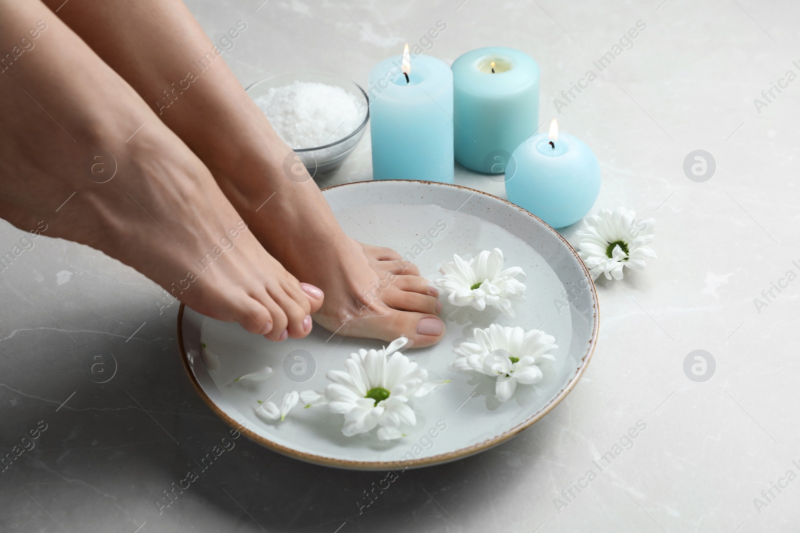 Photo of Woman soaking her feet in bowl with water and flowers on grey marble floor, closeup. Pedicure procedure