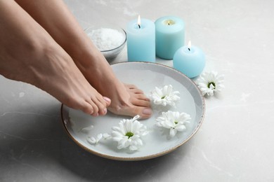 Woman soaking her feet in bowl with water and flowers on grey marble floor, closeup. Pedicure procedure