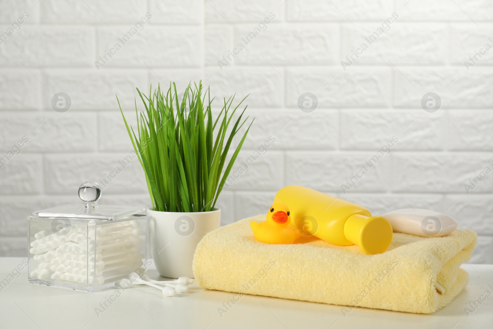 Photo of Baby bath accessories. Towel, cosmetic products, cotton swabs and toy duck on white table against brick wall