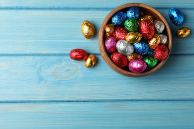 Bowl with chocolate eggs wrapped in colorful foil on light blue wooden table, flat lay. Space for text