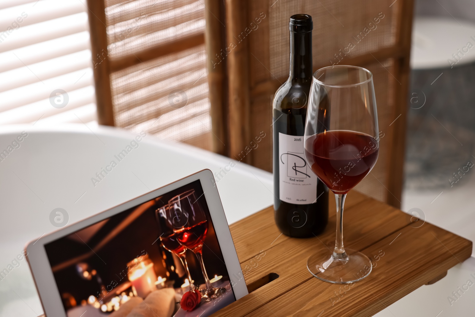 Photo of Wooden tray with tablet, glass of wine and bottle on bathtub in bathroom