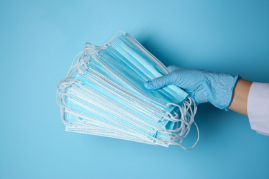 Photo of Doctor in latex gloves holding disposable face masks on light blue background, closeup. Protective measures during coronavirus quarantine