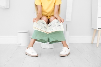 Young woman reading newspaper while sitting on toilet bowl at home