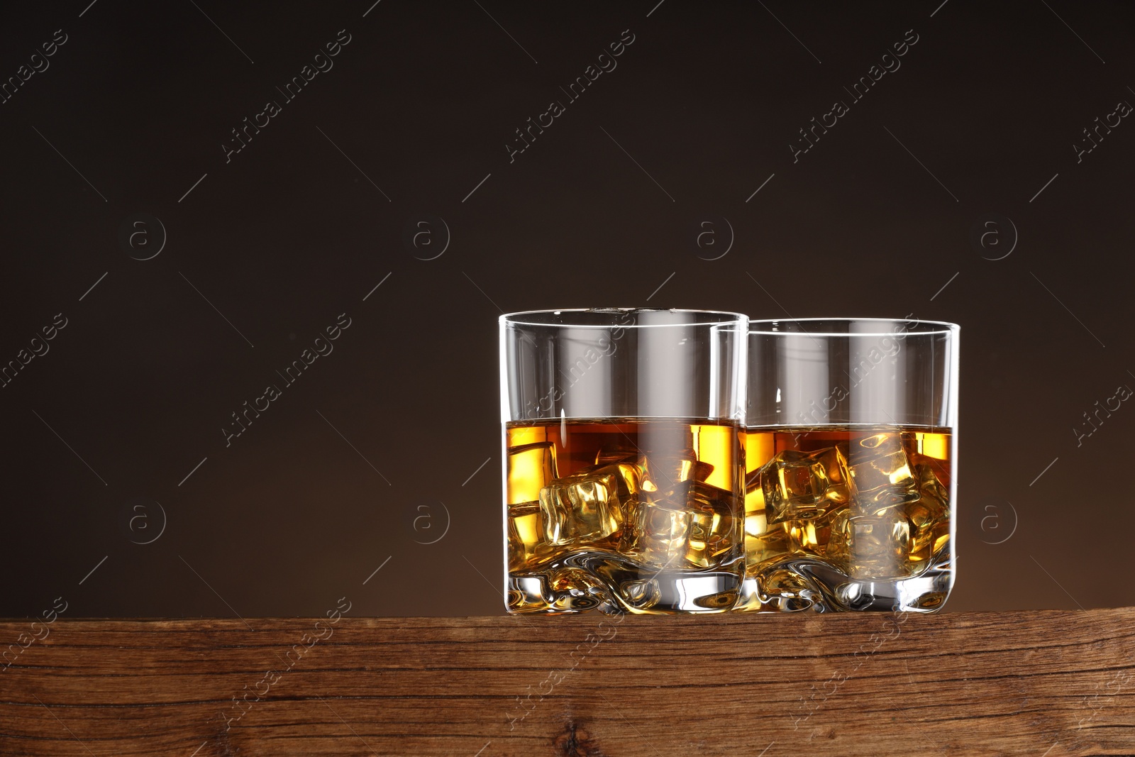 Photo of Whiskey with ice cubes in glasses on wooden table against brown background, space for text
