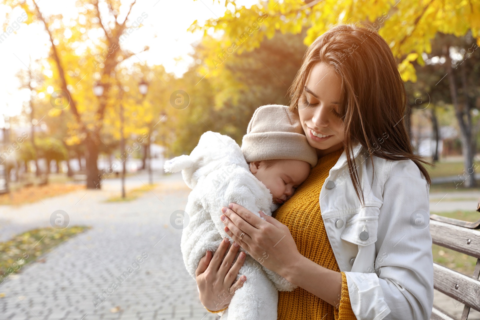 Photo of Happy mother with her sleeping baby son on bench in autumn park, space for text