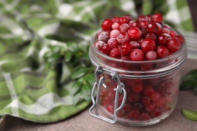 Frozen red cranberries in glass jar and green leaves on brown textured table, closeup. Space for text