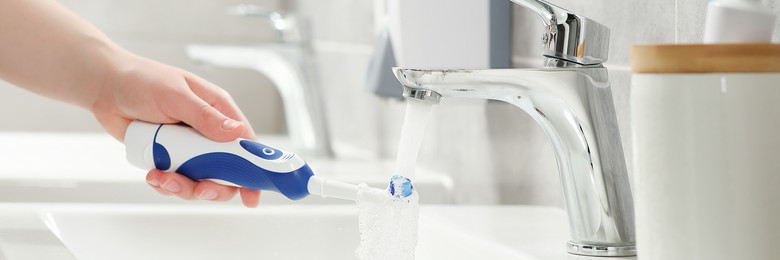 Image of Woman washing electric toothbrush under flowing water in bathroom, closeup. Banner design