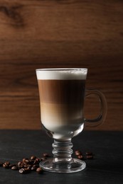 Glass cup of delicious layered coffee and beans on black table against wooden background, space for text