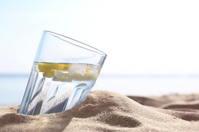 Photo of Sandy beach with glass of refreshing lemon drink on hot summer day, space for text