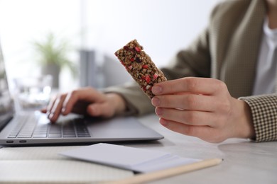 Woman holding tasty granola bar working with laptop at light table in office, closeup