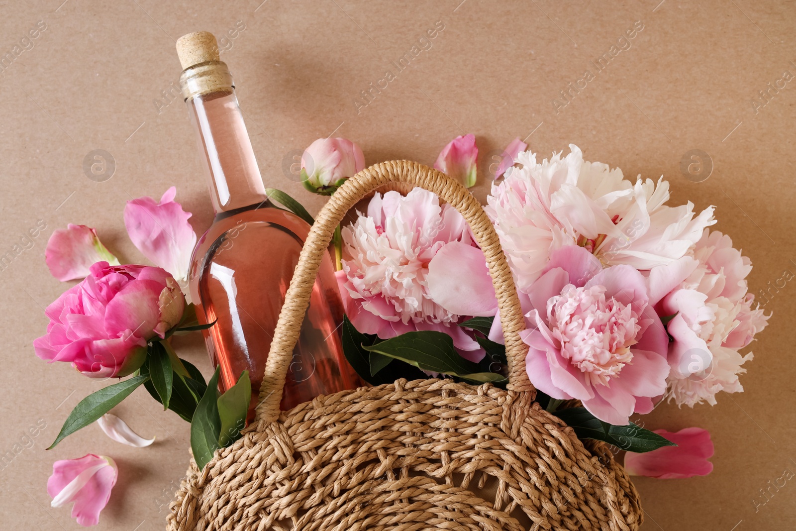 Photo of Wicker bag with bottle of rose wine and beautiful pink peonies on brown background, top view