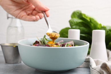 Photo of Woman pouring tasty vinegar based sauce (Vinaigrette) from spoon into bowl with salad at grey table, closeup