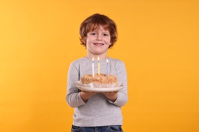 Photo of Birthday celebration. Cute little boy holding tasty cake with burning candles on orange background