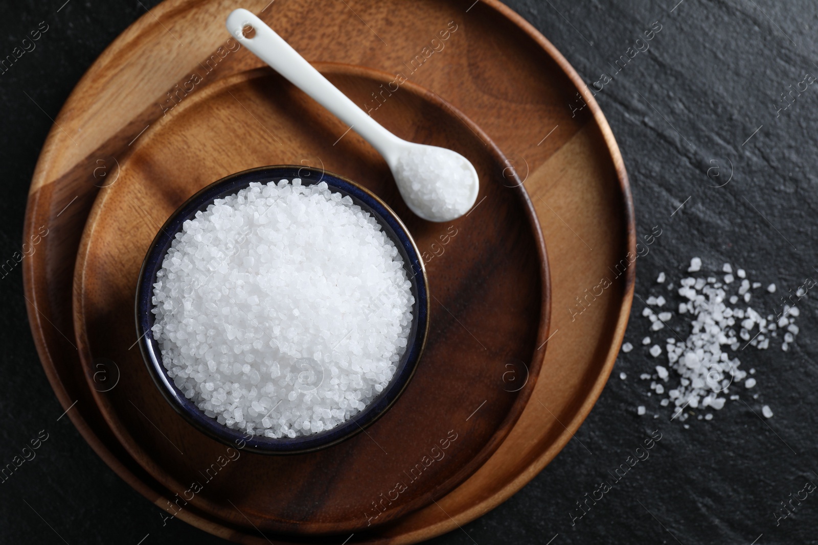 Photo of Organic white salt in bowl and spoon on black table, top view