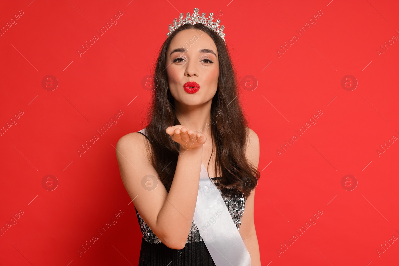 Photo of Beautiful young woman with tiara and ribbon in dress blowing kiss on red background. Beauty contest