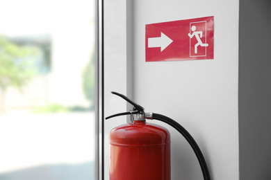 Photo of Modern fire extinguisher and emergency exit sign near window indoors