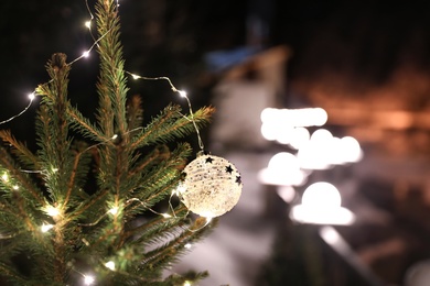 Beautiful conifer tree with glowing Christmas lights and ball on street, space for text. Winter holiday