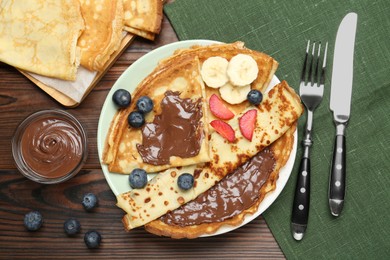 Photo of Tasty crepes with chocolate paste, banana and berries served on wooden table, flat lay