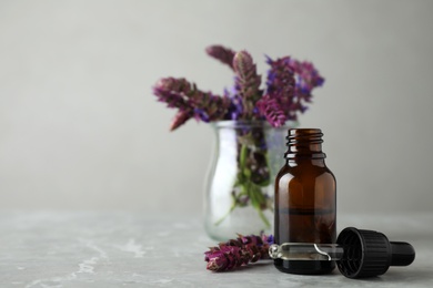 Photo of Bottle of sage essential oil and flowers on grey table, space for text