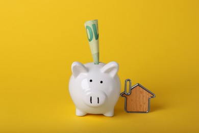 Photo of Ceramic piggy bank, euro banknote and key trinket on yellow background. Financial savings