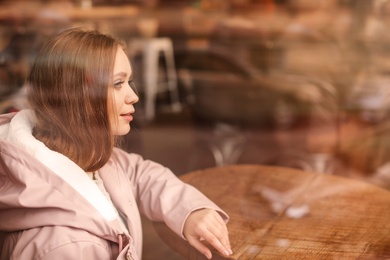 Photo of Beautiful young woman sitting at table in cafe, view from outdoors through window