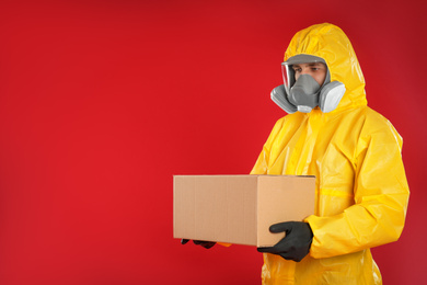 Photo of Man wearing chemical protective suit with cardboard box on red background, space for text. Prevention of virus spread