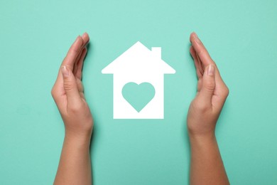 Image of Woman holding hands around paper silhouette of house on turquoise background, top view. Insurance concept