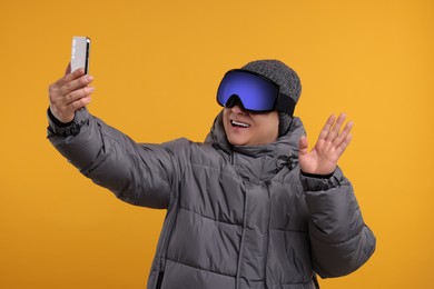 Winter sports. Happy man in ski goggles taking selfie on orange background