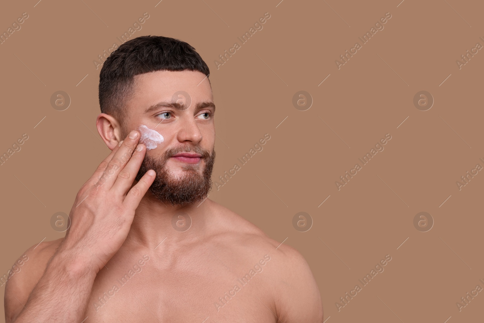 Photo of Handsome man applying cream onto his face on pale brown background, space for text