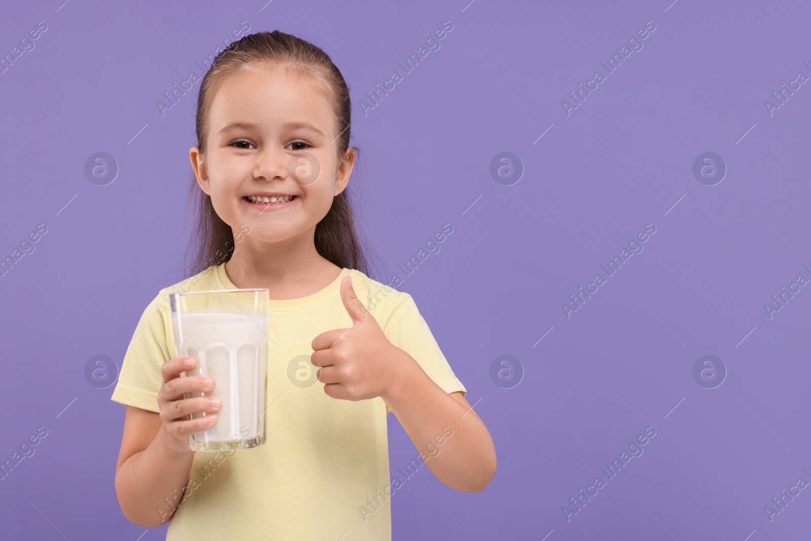 Photo of Cute girl with glass of fresh milk showing thumb up on violet background, space for text