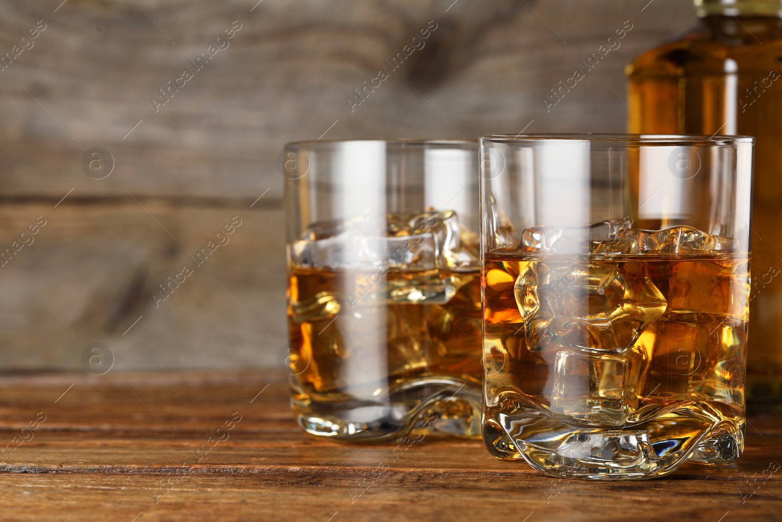 Photo of Whiskey with ice cubes in glasses and bottle on wooden table, closeup. Space for text