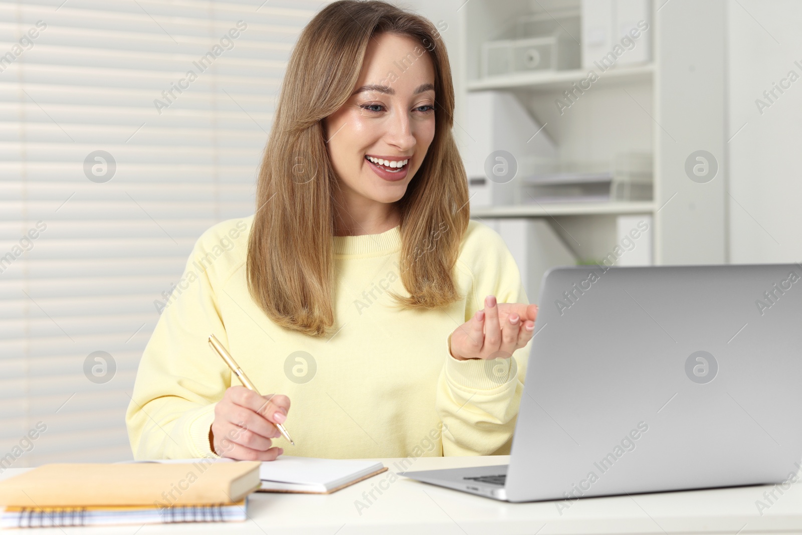 Photo of Young woman waving hello during video chat via laptop at white table indoors