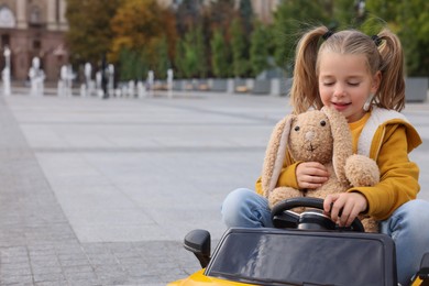 Photo of Cute little girl with toy bunny driving children's car on city street. Space for text