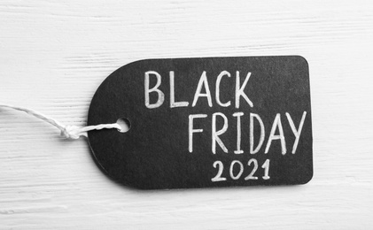 Photo of Tag with words BLACK FRIDAY 2021 on white wooden background, top view