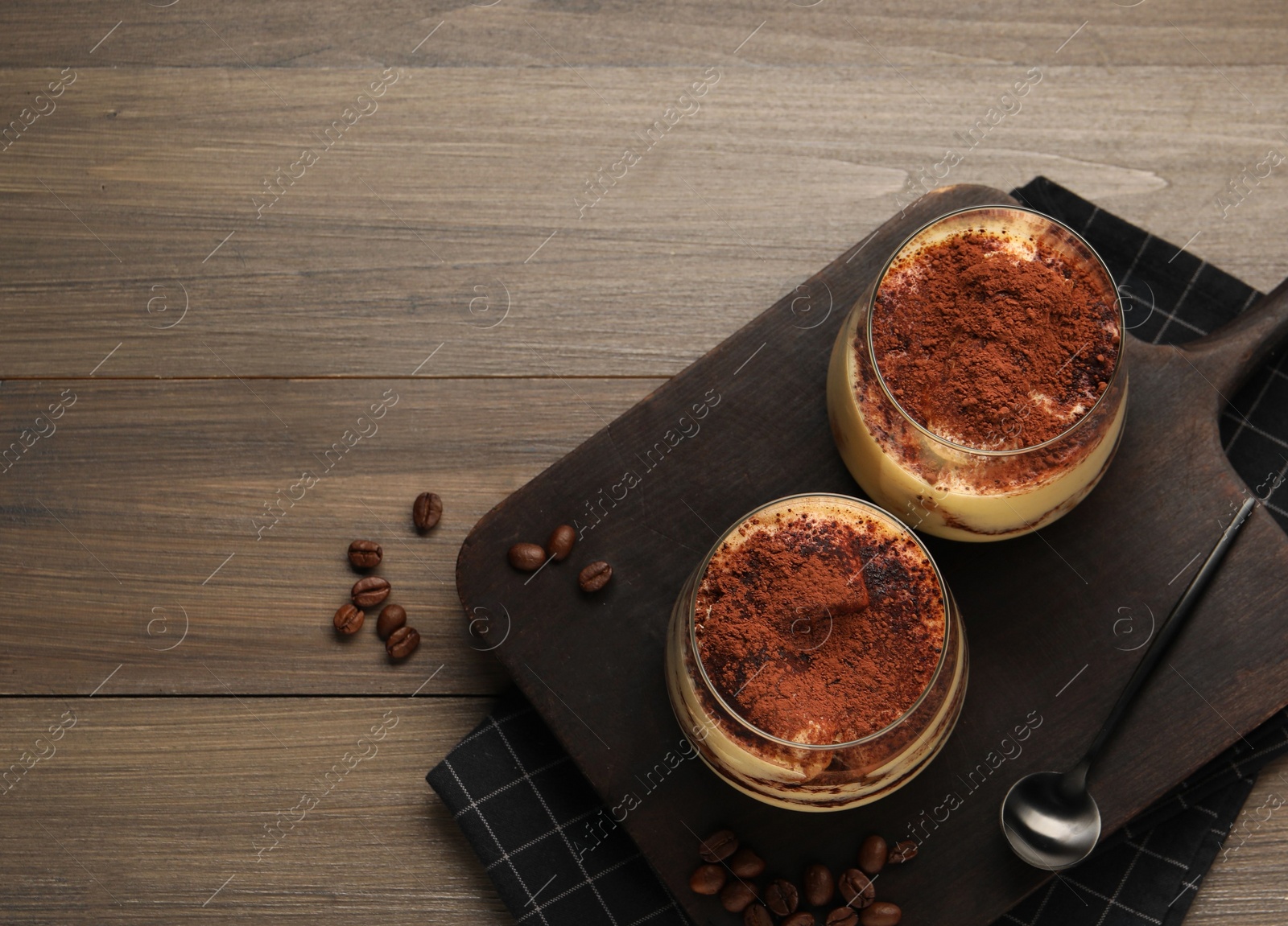 Photo of Delicious tiramisu in glasses, spoon and coffee beans on wooden table, top view. Space for text