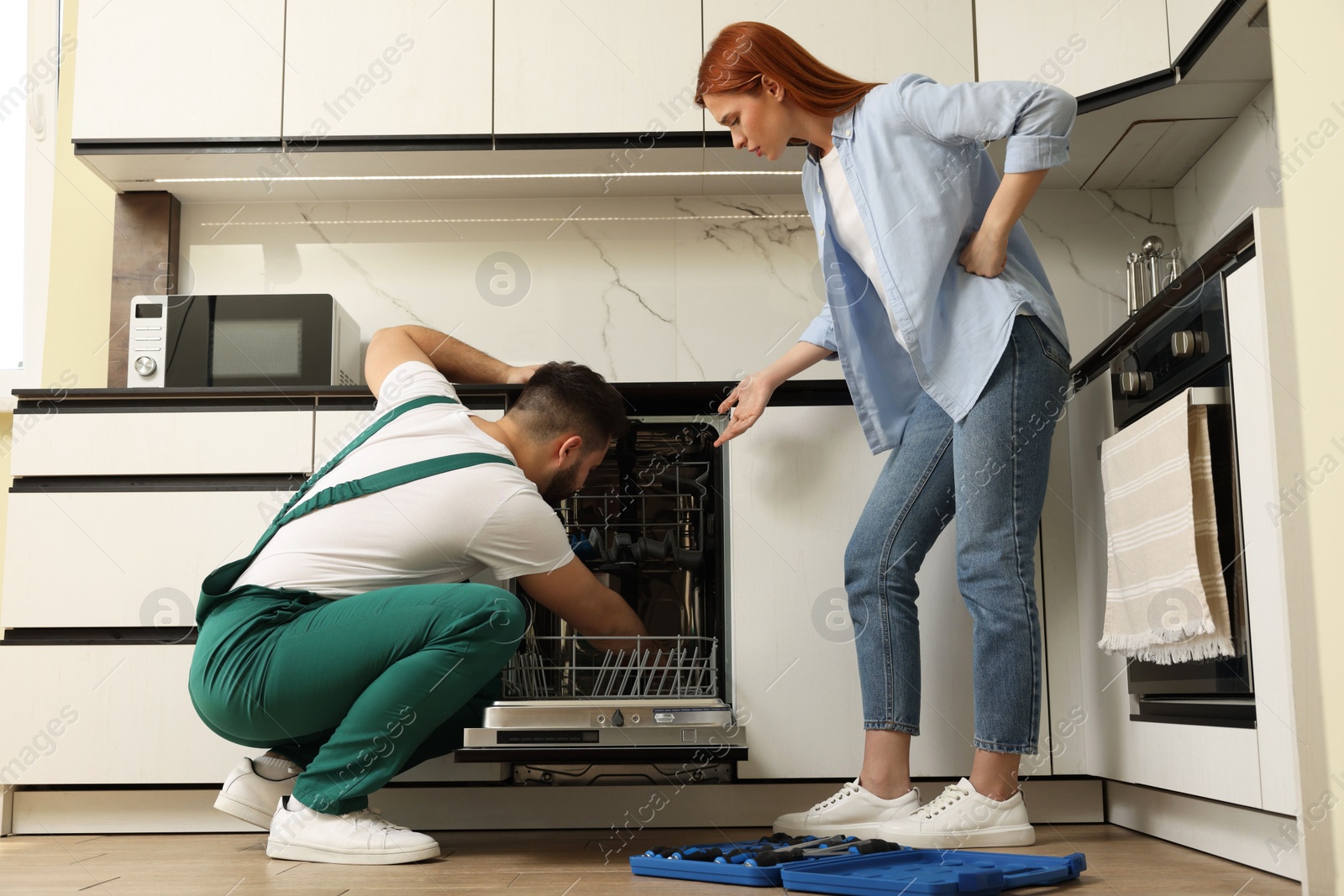 Photo of Young woman discussing with repairman near dishwasher in kitchen, low angle view